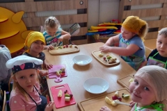 Grupa dzieci; 1 chłopiec i 5 dziewczynek ubrana na kolorowo; w fartuszkach i w nakryciach na głowę. Przed każdym dzieckiem leży deska do krojenia , a na niej owoce . Dzieci kroją na deskach owoce. Na środku stolika leżą 2 białe talerze. Za dziećmi widać 2 koloryfery w obudowie, pojemnik z torowiskiem i okna , a na nich kwiaty i wiosenne dekoracje.