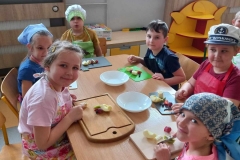 Grupa dzieci; 3 chłopców i 3 dziewczynki ubrana na kolorowo; w fartuszkach i w nakryciach na głowę. Przed każdym dzieckiem leży deska do krojenia , a na niej owoce . Dzieci kroją na deskach owoce. Na środku stolika leżą 2 białe talerze. Za dziećmi widać 2 koloryfery w obudowie, prostokątna szafka przedszkolna, szafka w kształcie słonka . Na parapetach okna dekoracje wiosenne.