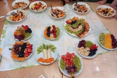 Na prostokątnym stoliku serwetka ,a na niej ułożone talerze z owocową sałatką i ciekawymi kompozycjami z owoców; palmy , delfinki, kolorowe tęcze, jeże.