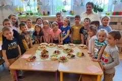 Grupa dzieci ubrana na kolorowo i dorosła kobieta zgromadzone wokół stolika na którym znajdują się na talerzach owocowe sałatki i ciekawe owocowe kompozycje; palmy , delfinki, kolorowe tęcze, jeże. Za grupą w tle okna z wiosenną dekoracją.