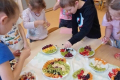 Kilka dzieci zgromadzonych wokół stolika degustuje przygotowane owocowe pyszności.. W tle mebelki przedszkolne i chłopiec bawiący się w kąciku kuchennym.