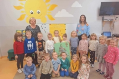 11 dzieci stoi w półkolu. 2 dziewczynki trzymają w rękach zielony karton. Na nim narysowana piramida i naklejone kolorowe obrazki. Przed grupą klęczy 6 dzieci. Za grupą stoją 2 panie. Na ścianie namalowane żółte słońce z promykami i 2 białe chmury.