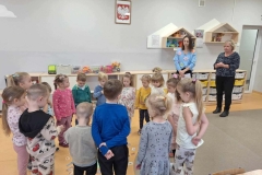 Dzieci stoją w kole. Obok nich stoją 2 panie w czarnych spodniach. Jedna ma niebieski swetr z kolorowymi kwiatami,  druga czarno- popielatą bluzkę. Za nimi stoi szafka z szufladami. Na ścianie wisi godło i 2 półki z kolorowymi maskotkami. Pomiędzy nimi kalendarz.