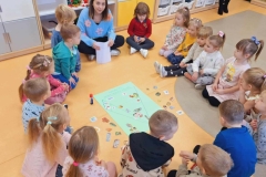 Grupa dzieci siedzi w kole na szarej i żółtej podłodze. Wśród nich pani w niebieskim swetrze z kolorowymi kwiatami. Na podłodze leży zielony karton z narysowaną piramidą i naklejonymi obrazkami. Obok porozrzucane ilustracje z produktami spożywczymi. Obok kartonu stoi klej z czerwoną zakrętką. Wysłano 11 dzieci stoi w pó