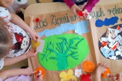 Na Zielonej Ziemi widać drzewo z bibuły, dziewczynki doklejają kolorowe kwiaty. Z boku widać część smutnej Ziemi na której naklejone są śmieci.