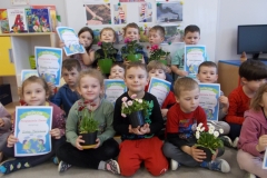 Dzieci przedszkolne ubrane w kolorowe stroje siedzą na podłodze w sali przedszkolnej. Trójka dzieci z przodu trzyma doniczki z kolorowymi stokrotkami, troje z tyłu trzyma doniczki z różowymi pelargoniami.Pozostałe dzieci mają w rękach niebieskie dyplomy z białą otoczką. Na dyplomach widoczny napis: Dyplom Przyjaciela Ziemi otrzymuje ( imię i nazwisko dziecka).Pod spodem rysunek chłopca i dziewczynki obejmujących kulę ziemską. W tle widoczny kącik książki, tablica tematyczna i kącik komputerowy.