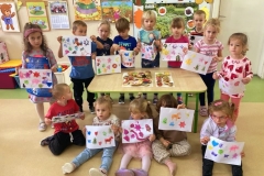 Grupa dzieci ustawiona wokół stolika, piecioro z nich siedzi na podłodze przed stolikem, trzymają w rękach kolorowe prace, na stoliku leżą stemple wykonane z ziemniaków.