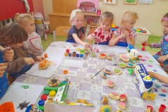 Grupa dzieci siedzi przy stoliku na którym leżą kolorowe farby, stemple wykonane z ziemniaków. Dzieci malują stemple farbami i odbijają na kartce.