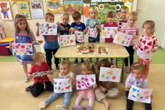 Grupa dzieci stoi wokół stołu, pięcioro z nich siedzi na podłodze przed stolikiem, w rękach trzymają kolorowe prace, na stoliku leżą stemple wykonane z ziemniaków.