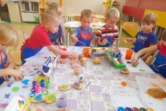 Grupa dzieci siedzi przy stoliku na którym leżą kolorowe farby, stemple wykonane z ziemniaków. Dzieci malują stemple farbami i odbijają na kartce.