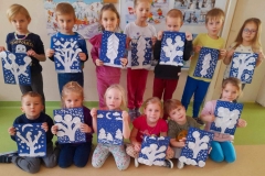 Kilkoro dzieci stoi przed tablicą, kilkoro siedzi przed nimi na podłodze, każde z nich trzyma w ręku zimowa pracę.