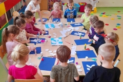 Dzieci siedzą przy stolikach, kleją na granatowej kartce zimowe drzewa z płatków kosmetycznych.