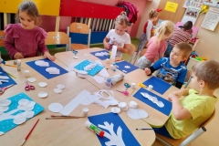 Kilkoro dzieci siedzi przy stolikach, na granatowa kartkę naklejają białe elementy, tak by powstał zimowy krajobraz.