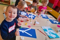 Grupa dzieci siedzi przy stolikach, naklejają na granatowa kartkę płatki kosmetyczne.