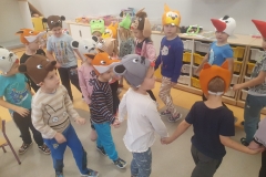 Sala zabaw . Dzieci ubrane na kolorowo z maskami zwierząt na głowach trzymają się za ręce i poruszają po obwodzie koła w rytm muzyki.