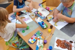 Troje dzieci; chłopiec i dwie dziewczynki siedzą przy prostokątnym stoliku i malują farbami kontury zwierząt na białych kartkach . Dzieci mają założone fartuszki do malowania . Na stole rozłożone pojemniki z farbami i na wodę.