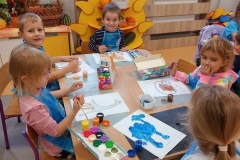 Pięcioro dzieci; dwóch chłopców i trzy dziewczynki siedzą przy prostokątnym stoliku i malują farbami kontury zwierząt na białych kartkach . Dzieci mają założone fartuszki do malowania . Na stole rozłożone pojemniki z farbami i na wodę. Za dziećmi w tle kącik przyrody z darami jesieni . Powyżej widać fragment okna . Na oknie wizytówki z imionami dzieci i dwa kwiaty doniczkowe. Pod oknem kaloryfer zabezpieczony meblową drabinką.