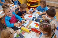 Sześcioro dzieci; dwójka chłopców i cztery dziewczynki siedzą przy prostokątnym stoliku i malują farbami kontury zwierząt na białych kartkach . Dzieci mają założone fartuszki do malowania . Na stole rozłożone pojemniki z farbami i na wodę. Za dziećmi w tle widać kącik przyrody z darami jesieni . Na oknie wizytówki z imionami dzieci . Pod oknem kaloryfer zabezpieczony meblową drabinką. Z lewej strony widać część stolika i dwóch chłopców ;jeden stoi i maluje , drugi z głową zwróconą w prawą stronę patrzy na obrazek kolegi.