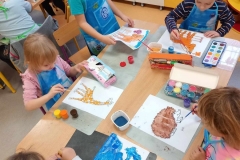 Piątka dzieci ; dwójka chłopców i trzy dziewczynki siedzą przy prostokątnym stoliku i malują farbami kontury zwierząt na białych kartkach . Jedem chłopiec stoi podczas malowania. Dzieci mają założone fartuszki do malowania . Na stole rozłożone pojemniki z farbami i na wodę. Za dziećmi w tle kącik przyrody z darami jesieni . Z lewej strony widać część stolika i dziewczynkę malującą farbami