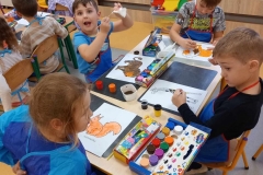 Czwórka dzieci ; trzech chłopców i jedna dziewczynką siedzą przy prostokątnym stoliku i malują farbami kontury zwierząt na białych kartkach . Jeden z chłopców wyciera pędzelek robiąc przy tym zabawną minę. Dzieci mają założone fartuszki do malowania . Na stole rozłożone pojemniki z farbami i na wodę. Za dziećmi widać dwa kaloryfery zabezpieczone meblową drabinką.