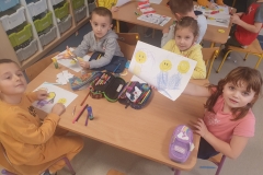 Przy pierwszym stoliku w sali przedszkolnej siedzi dwóch chłopców i dwie dziewczynki. Jedna z nich prezentuje swoją pracę plastyczną. Są na niej trzy wycięte żółte uśmiechnięte głowy emotki i dorysowane na niej niebieskie ubrania. Przed pozostałymi dziećmi leżą ich rysunki. Dzieci wycinają, przyklejają i rysują. Na stoliku leżą też piórniki, kleje i pocięty papier. W tle znajduje się drugi stolik z pięciorgiem tylko częściowo widocznych dzieci. One też wykonują prace plastyczne.
