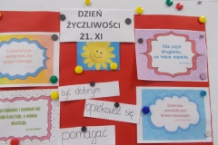 Na białej tablicy przypięty jest czerwony karton. U góry na nim widoczny jest czarny drukowany napis na białym tle: Dzień Życzliwości  21. XI. Pod nim jest żółte uśmiechnięte słoneczko na niebieskim tle. Pod słoneczkiem znajdują się trzy odręczne napisy na białych, wąskich paskach papieru: być dobrym, opiekować się, pomagać. Po obu stronach kartonu przypięte są magnesami przysłowia w ozdobnych ramkach o treściach: Nie czyń drugiemu, co tobie niemiłe, Dzień bez uśmiechu jest  dniem straconym, Człowiek jest warty tyle, ile uczyni dobrego, Uprzejmość i uśmiech nic nie kosztują a wielce skutkują.