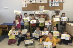 W sali przedszkolnej stoją i siedzą w gromadce dzieci w kolorowych strojach, przeważa w nich kolor żółty. Prezentują wykonane przez siebie prace plastyczne ,, W krainie Życzliwków’’. Do wyciętych uśmiechniętych żółtych twarzy - emotek dorysowywały elementy ciała i ubrań. W tle widoczne są tablice korkowe, na nich przypięte plansze z literami i cyframi.