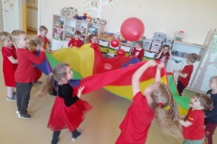 Dzieci podrzucają do góry czerwone balony ułożone na chuście animacyjnej. Dzieci są wesołe, radosne.