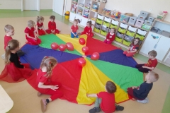 Dzieci ubrane na czerwono siedzą na chuście animacyjnej, na środku chusty rozłożone czerwone balony.
