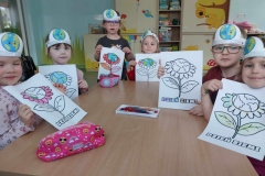 Dzieci z opaskami na głowie siedzą przy brązowym stoliku. W rękach trzymają rysunki z kwiatami. Na stoliku leży kolorowy piórnik.