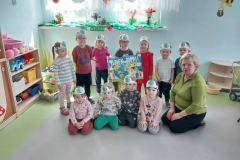 Grupa dzieci stoi w półkolu na szarej podłodze. Przed nimi siedzi czworo dzieci i pani w zielonym swetrze. Dzieci mają na głowie opaski z obrazkiem Ziemi. 2 dziewczynki trzymają plakat z napisem Dzień Ziemi. Za nimi na oknie kolorowe kwiaty.