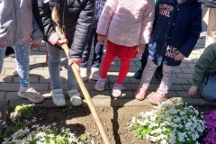 Dziewczynka w ciemnej kurtce, jasnych spodniach i czapce na głowie trzyma w rękach motykę. Robi nią w ziemi dołek. Obok niej stoją dziewczynki. Wokół kolorowe kwiaty.