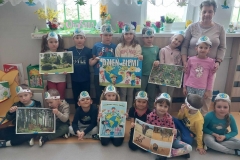 Dzieci z opaskami na głowie stoją na tle okna. Przy nich pani w beżowym swetrze. Pozostałe dzieci siedzą na podłodze. 2 dzieci trzyma plakat z napisem Dzień Ziemi. 5 dzieci ma obrazki przedstawiające przyrodę.