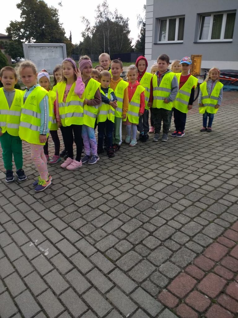 Dzieci ubrane w kamizelki odblaskowe ustawione parami stoją na chodniku. Ich głowy zwrócone są w lewą stronę. W tle widać szarą tablicę ogłoszeń i korony drzew. Obok część budynku w kolorze popielatym z 2 oknami
