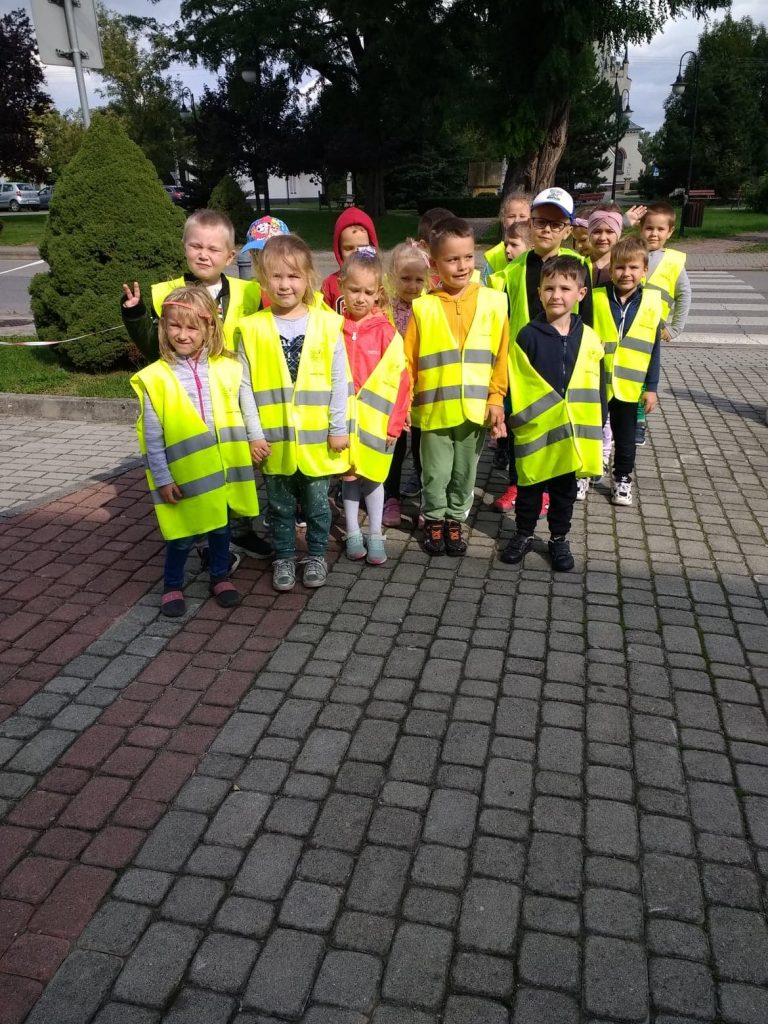 Grupa dzieci stoi na chodniku z kostki brukowej. Dzieci ubrane są w kamizelki odblaskowe. 3 chłopców ma na głowienka czapki, jedna dziewczynka opaskę. Wszystkie dzieci patrzą prosto przed siebie. Za nimi widać zielone krzewy