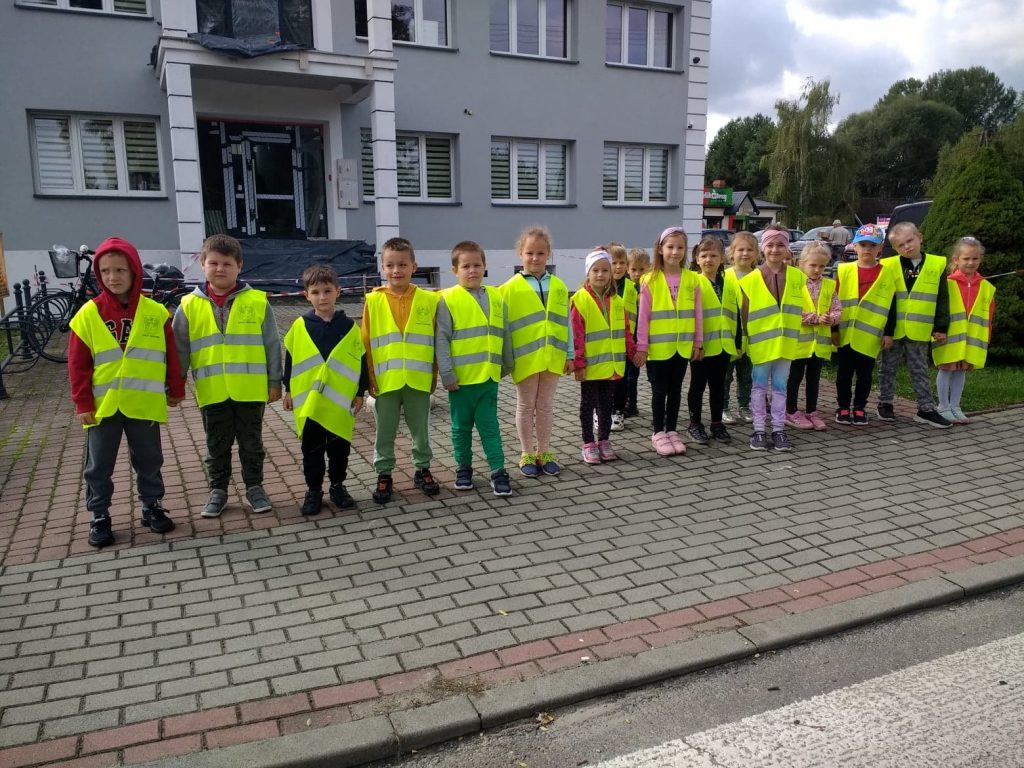 Grupa dzieci ustawiona w szeregu zwrócona jest przodem do ulicy. Przed nimi fragment przejścia dla pieszych. Za ich plecami szary budynek z oknami. 