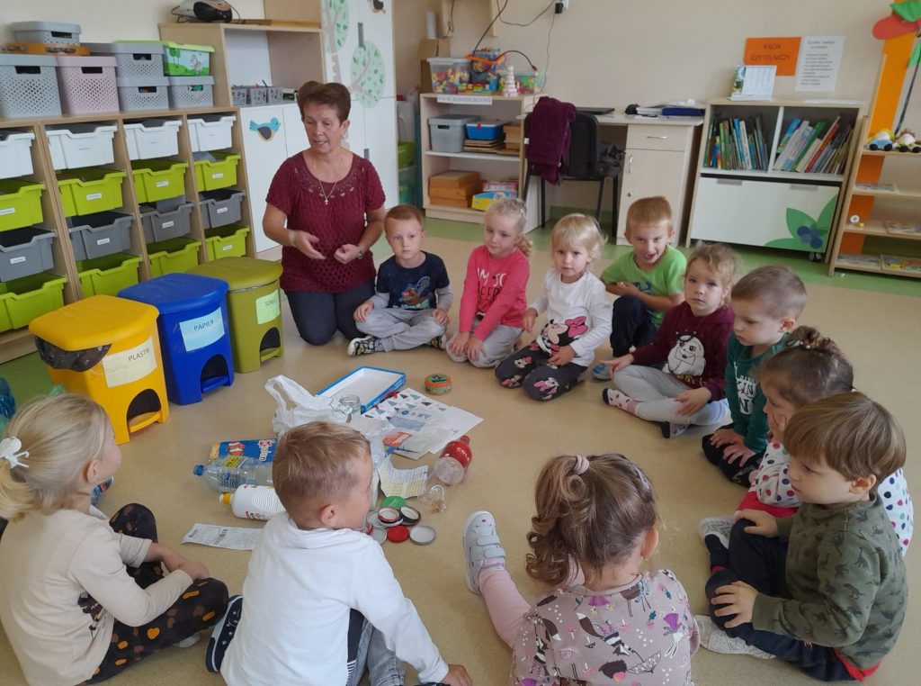 Grupa dzieci siedzi w kole zastanawia się do jakich pojemników wrzucić odpady