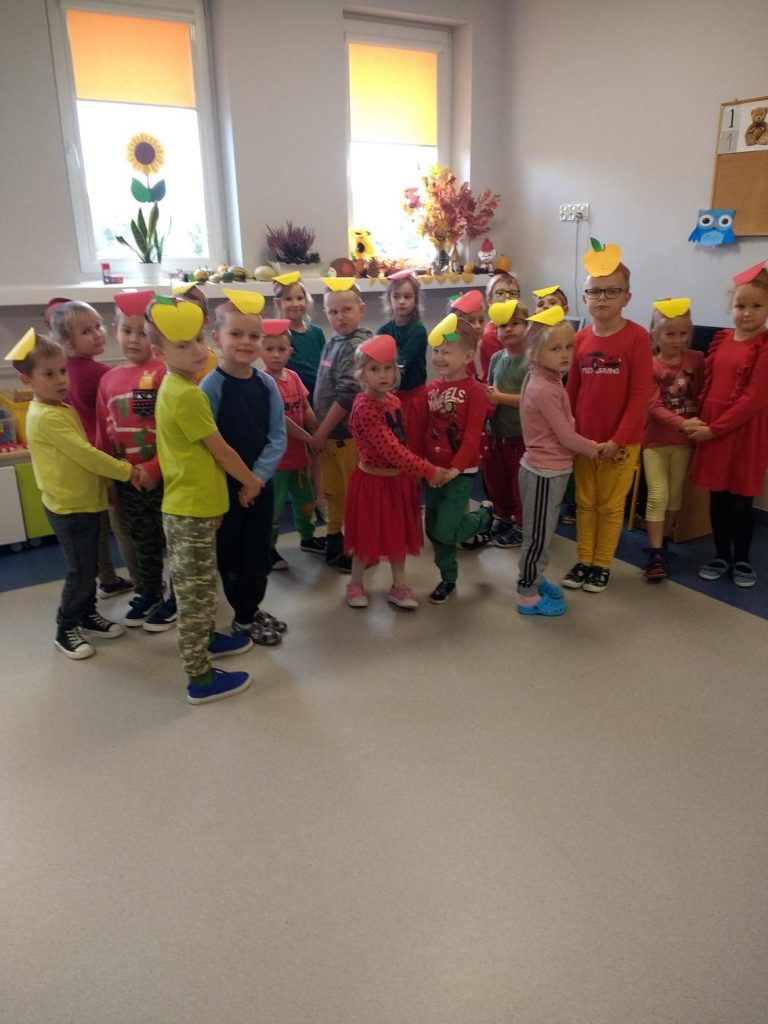 Dzieci w ubraniach głównie w kolorze czerwonym, zielonym i żółtym stoją w parach trzymając się za ręce. Na ich głowach opaski z czerwonym lub żółtym jabłkiem. Dzieci patrzą prosto przed siebie. Za nimi 2 okna z jesienną dekoracją.   