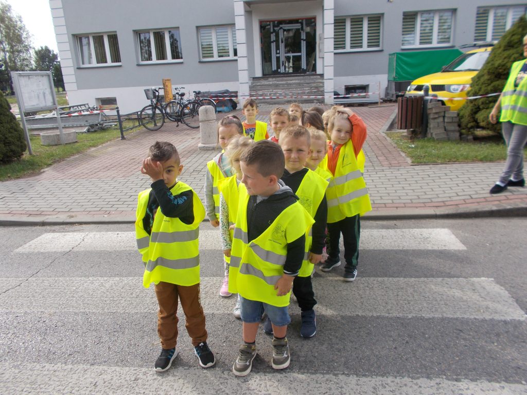 Ustawiona w pary grupa dzieci przedszkolnych, ubranych w żółto - srebrne kamizelki odblaskowe przechodzi po pasach na drugą stronę drogi. Z prawej strony częściowo widoczna opiekunka grupy. W tle budynek Urzędu Gminy zabezpieczony taśmami ze względu na remont. Przed nim po prawej stronie fragment zieleni, ułożona w stertę kostka brukowa, za nią żółty samochód i zielona kryta przyczepka samochodowa. Po lewej stronie stoją dwa rowery w stojaku, widoczna też jest tablica ogłoszeniowa.