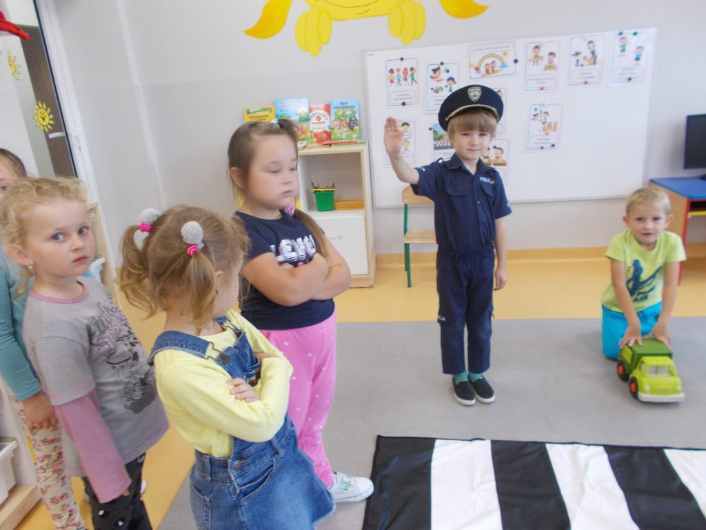 Czworo dzieci: dwóch chłopców i dwie dziewczynki stoją przed matą - przejściem dla pieszych. Z prawej strony stoi chłopiec w stroju policjanta, rękę ma uniesioną do góry. Obok niego klęczy dwóch chłopców, trzymających w rękach samochody. Z lewej strony widoczny chłopiec w ciemnej bluzie. Ma rękę podniesioną do góry. Obok niego stoi duży kolorowy samochód.