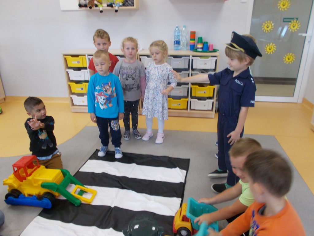 Mata imitująca przejście dla pieszych rozłożona jest w sali przedszkolnej. Z jej lewej strony stoją cztery dziewczynki ubrane w kolorowe stroje, czekają na możliwość przejścia. Ruchem kieruje stojący na środku chłopiec ubrany w strój policjanta. Prawą rękę ma uniesioną do góry. Obok niego klęczy chłopiec, ubrany w żółtą koszulkę i niebieskie spodnie. W ręce trzyma samochód. Z tyłu tablica z obrazkami oraz szafka z ustawionymi książkami.