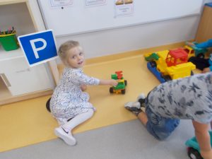 Miejsce w sali oznaczone plastikowym stojącym znakiem parkingu: niebieski kwadrat w białej ramie, na nim wielka biała drukowana litera P. Dziewczynka w jasnej sukience we wzorek wjeżdża  samochodem zabawką, aby zaparkować. Buzia odwrócona w kierunku osoby fotografującej. Z boku stoją dwa samochody. Chłopiec w koszulce z nadrukiem i w niebieskich spodniach odjeżdża z parkingu samochodem. Obok szafka, na półce kredki w pojemniku.