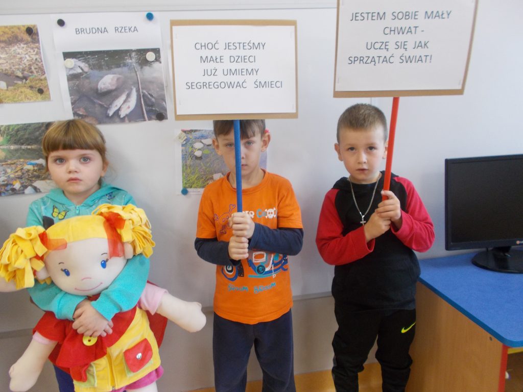 Troje dzieci stoi przed biała tablicą z planszami, których tematem są zanieczyszczone rzeki i śnięte ryby. Chłopiec w ciemno - szarym ubraniu i okularach trzyma przed sobą szmacianą lalkę chłopca w żółto - niebieskim stroju, z ciemnymi włosami. Dziewczynka ubrana w niebieską bluzę trzyma szmacianą lalkę - dziewczynkę, z jasnymi włosami, ubraną w czerwono - żółty strój. Obok stoi chłopiec w pomarańczowej bluzie z nadrukiem i ciemnych spodniach. Trzyma w ręku niebieski patyk do którego przypięta jest biała kartka z napisem drukowanymi literami: „Choć jesteśmy małe dzieci już umiemy segregować śmieci”.