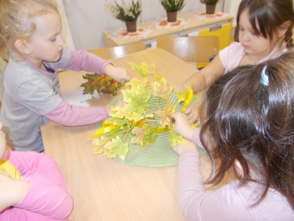 W sali przedszkolnej przy stoliku siedzą cztery dziewczynki, dwie z nich widoczne tylko częściowo ( z boku i tył głowy). Stroją one zielony kapelusz we wstążki i liście w kolorach żółtym, zielonym i brązowym. W tle widoczna szafka oraz wrzosy w doniczkach.
