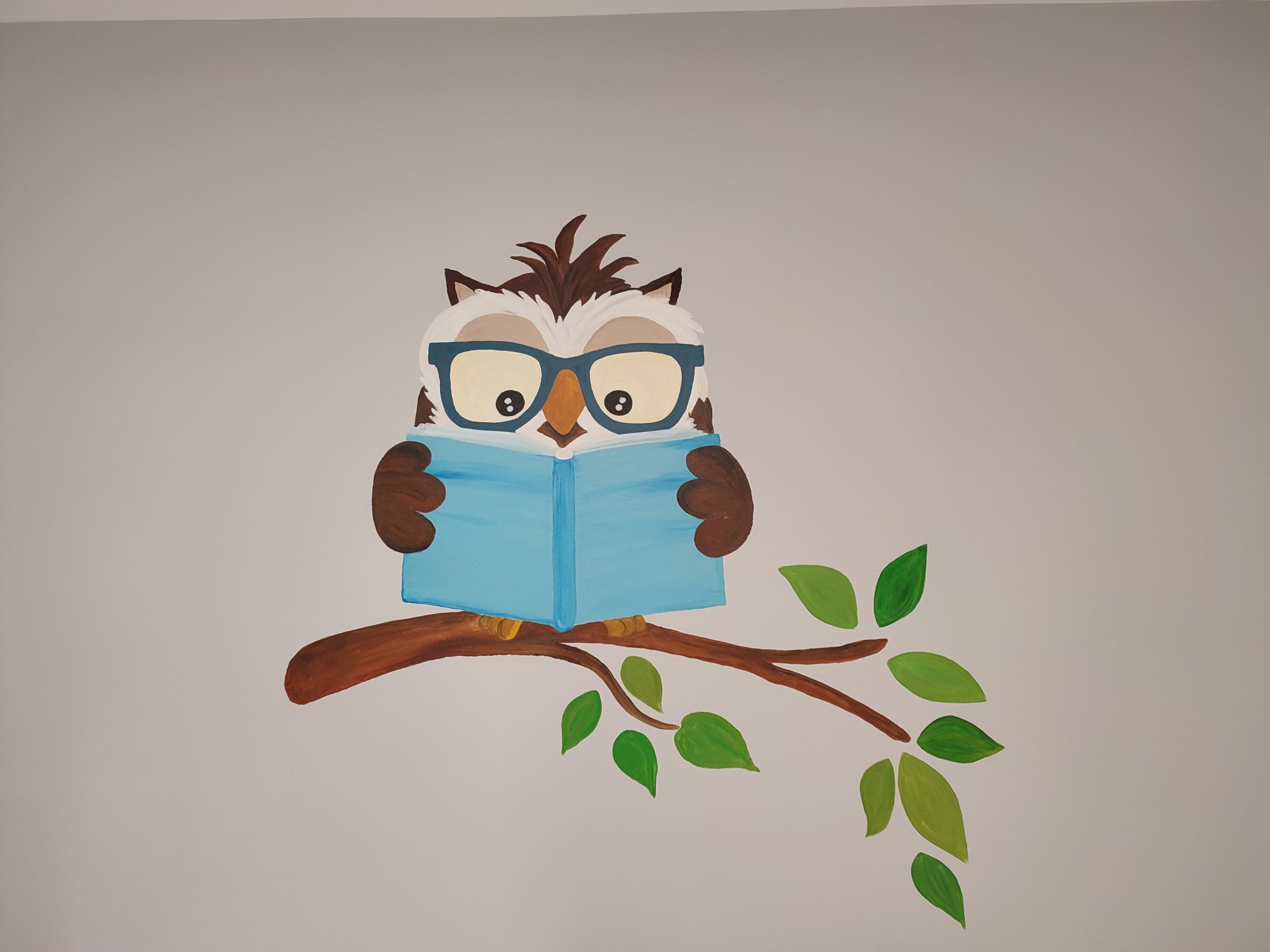 Namalowana na ścianie brązowa sowa w granatowych okularach, trzyma niebieską książkę, siedzi na gałązce z zielonymi listkami