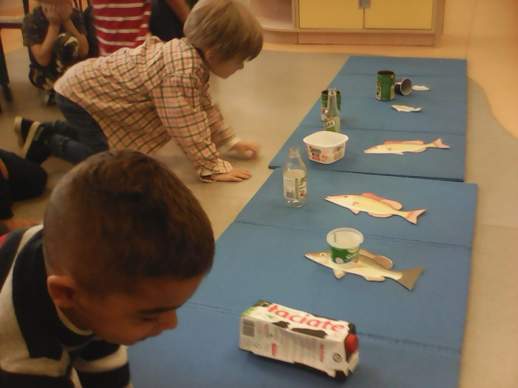 Na podłodze sali ułożone dwie wąskie niebieskie maty piankowe obrazujące rzekę. Na nich znajdują się wycięte z kartonu sylwety ryb oraz szklane butelki, puszki, pojemniki plastikowe oraz karton po mleku. ż lewej strony dwóch chłopców nachyla się nad matą. Pierwszy ubrany w biało - czarną bluzę, drugi w jasną koszulę w kratkę.