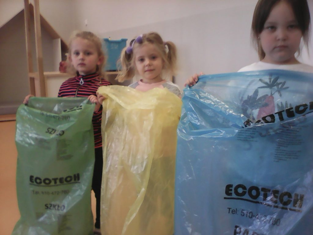 Trzy dziewczynki, u których widoczne są tylko głowy i górne fragmenty odzieży stoją obok siebie w sali przedszkolnej. Przed sobą trzymają worki na odpady w kolorach zielonym, żółtym i niebieskim. Na workach widoczne są napisy czarnymi, drukowanymi literami o treści: Ecotech.
