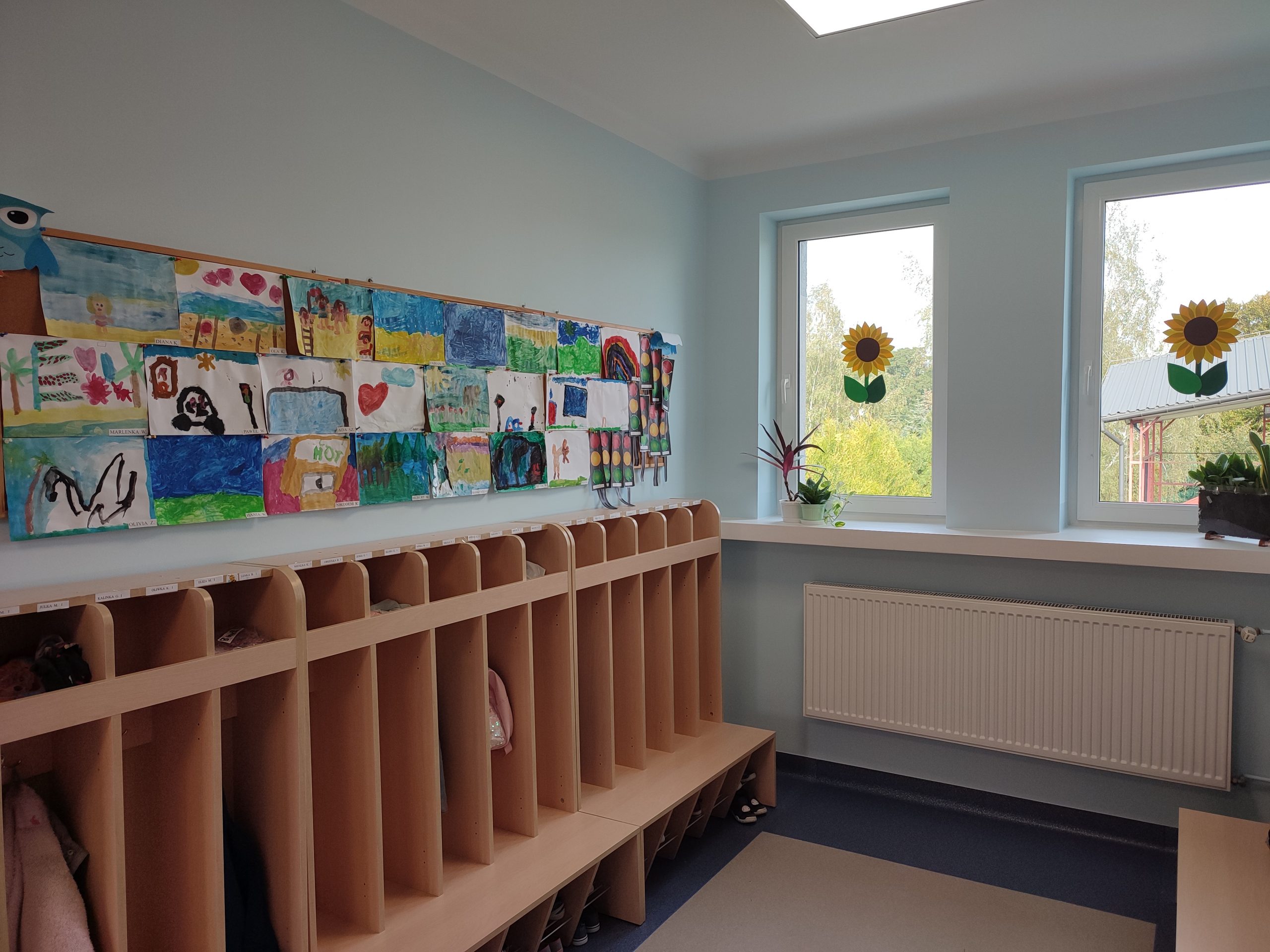 szafki szatniowe z przegródkami w jasnym kolorze drewna, nad nimi kolorowe prace dzieci wykonane na kartkach papieru, po lewej stronie okno z przyklejonym na szybie słonecznikiem, na parapecie kwiatek doniczkowy