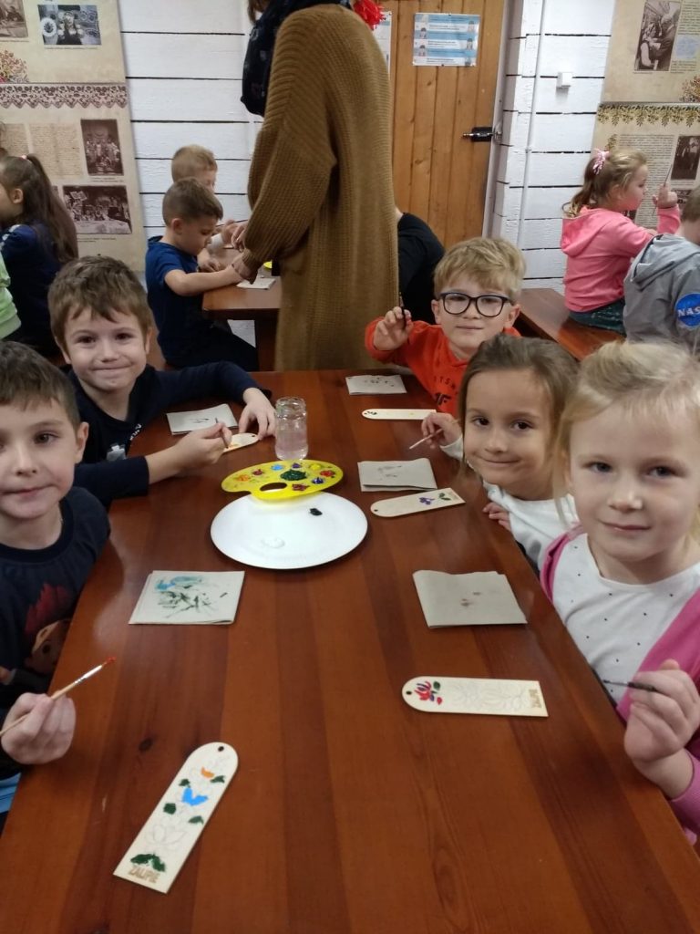 Dzieci siedzą przy stolikach. W rękach trzymają pędzelki. Przed nimi drewniane zakładki ze wzorem do malowania. Na stołach farby i słoiki z wodą oraz papier do wycierania pędzla.