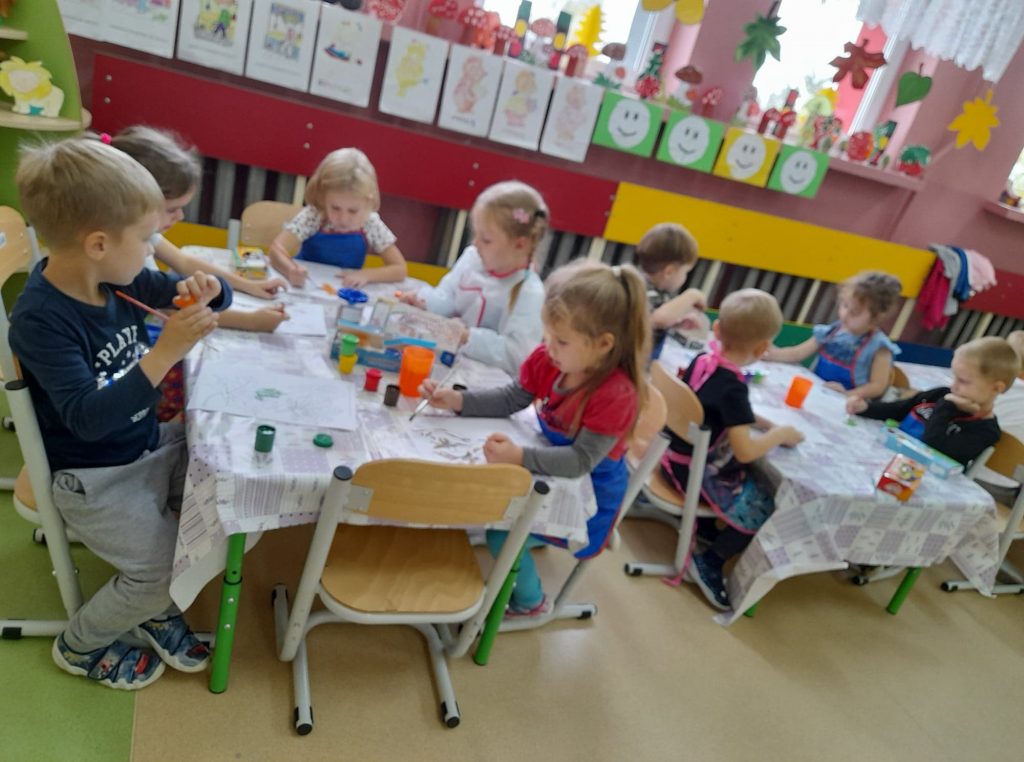Na zdjęciu widać dwa stoliki przy których siedzą dzieci. Dzieci ubrane są w kolorowe fartuszki , malują farbami. Na stole rozłożone są kolorowe farby. W tle Na parapetach widać kolorowe rysunki dzieci.